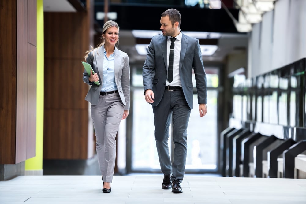 Two professional people walking and discussing business in an office entry way with large windows behind them